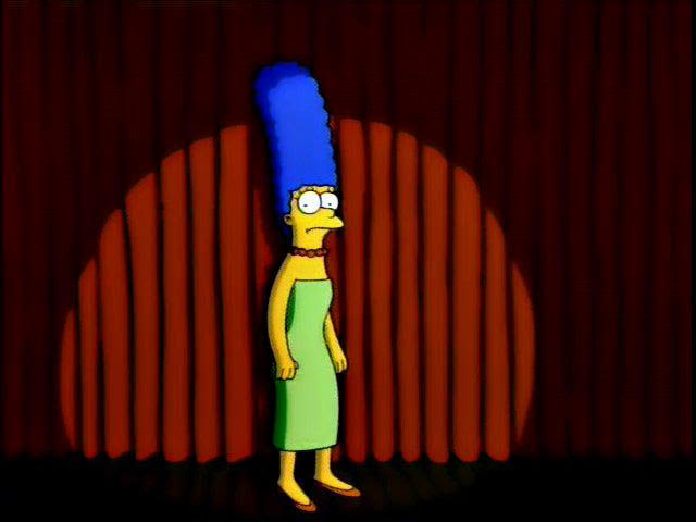 Marge Simpson on stage, under a spotlight, a red curtain behind her