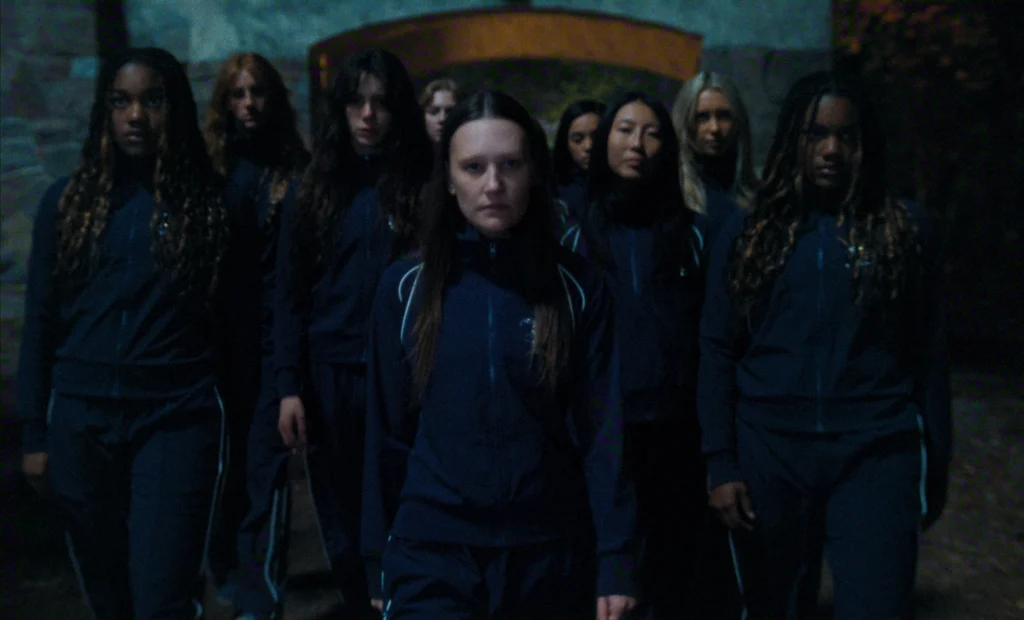 "A group of serious-faced young women in matching navy tracksuits stand outdoors in low light, possibly at night, in front of a stone archway surrounded by trees."