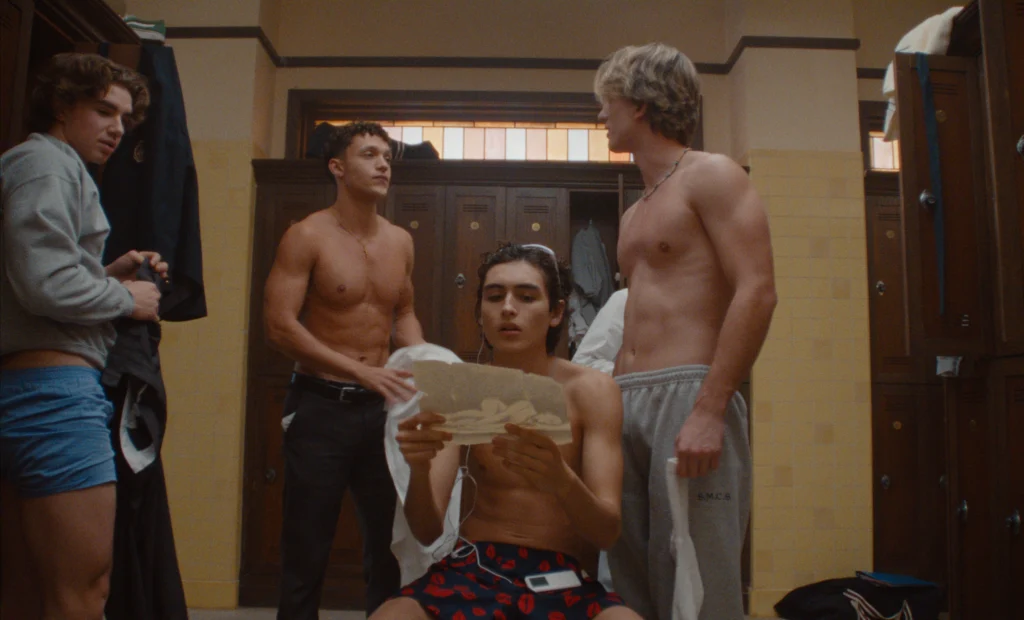 "Four young men in a locker room, some shirtless, one sitting on a bench reading a letter while the others stand around him with lockers and gym bags in the background. The setting has yellow tile walls and a relaxed atmosphere."