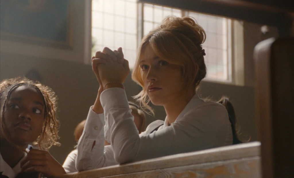 "Two young women sit in a sunlit church pew, one looking ahead with hands clasped in prayer, the other glancing sideways; large windows illuminate the warm, calm interior setting."