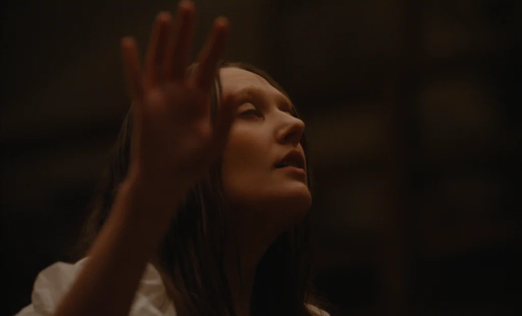 "A person with long hair, eyes closed, raises their hand in a dimly lit indoor environment, possibly expressing emotion or concentration, with a soft brown background."