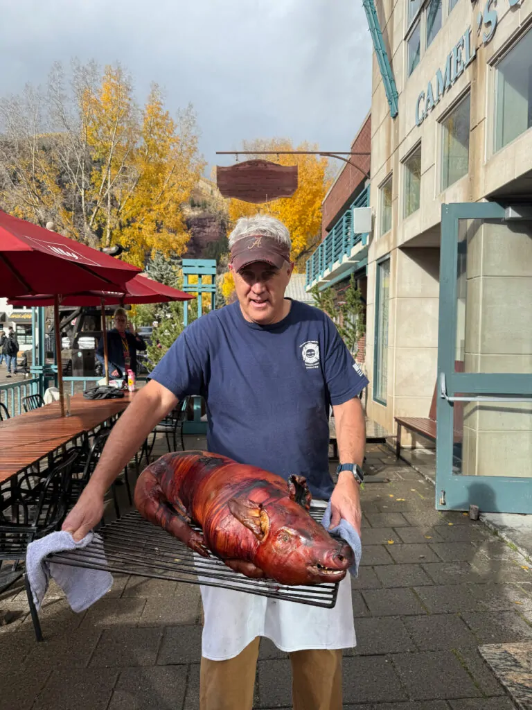 A man holding a roast pig outside, with tented tables and quaking aspens behind him