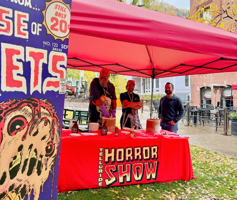 Three men stand under a tent at a table with a red cloth with the Telluride Horror Show logo. They're serving ice cream and smiling