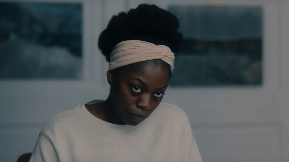 A still from Joan Iyiola's MANGO. A Black woman with her hair up wearing a white hairband looks seriously just past the camera.