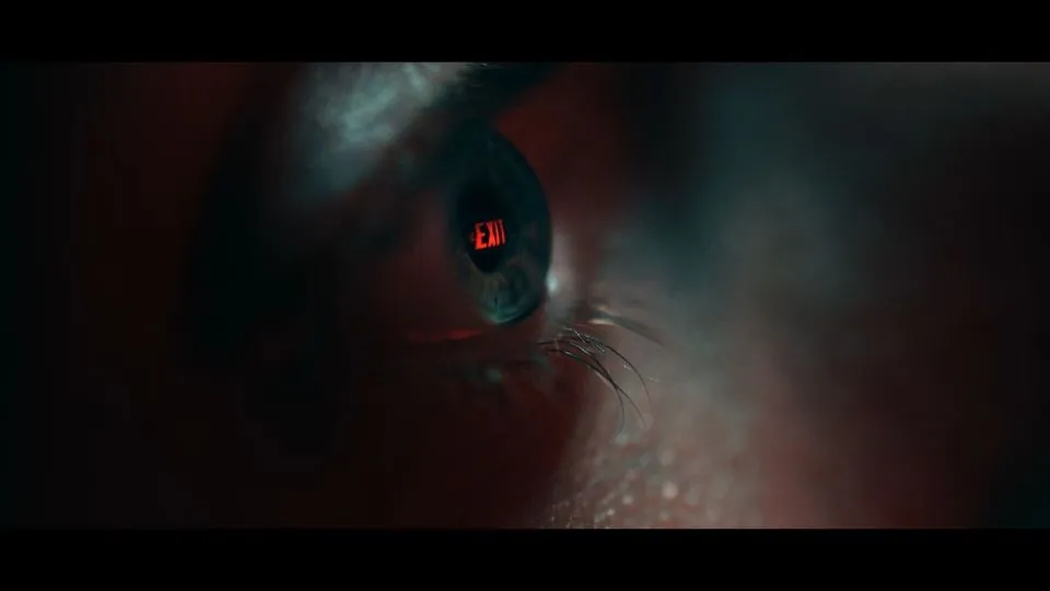 A still from Greg Jeffs's TRAPPED. A close up of a white man's blue eye. A red exit sign is reflected in the eye.