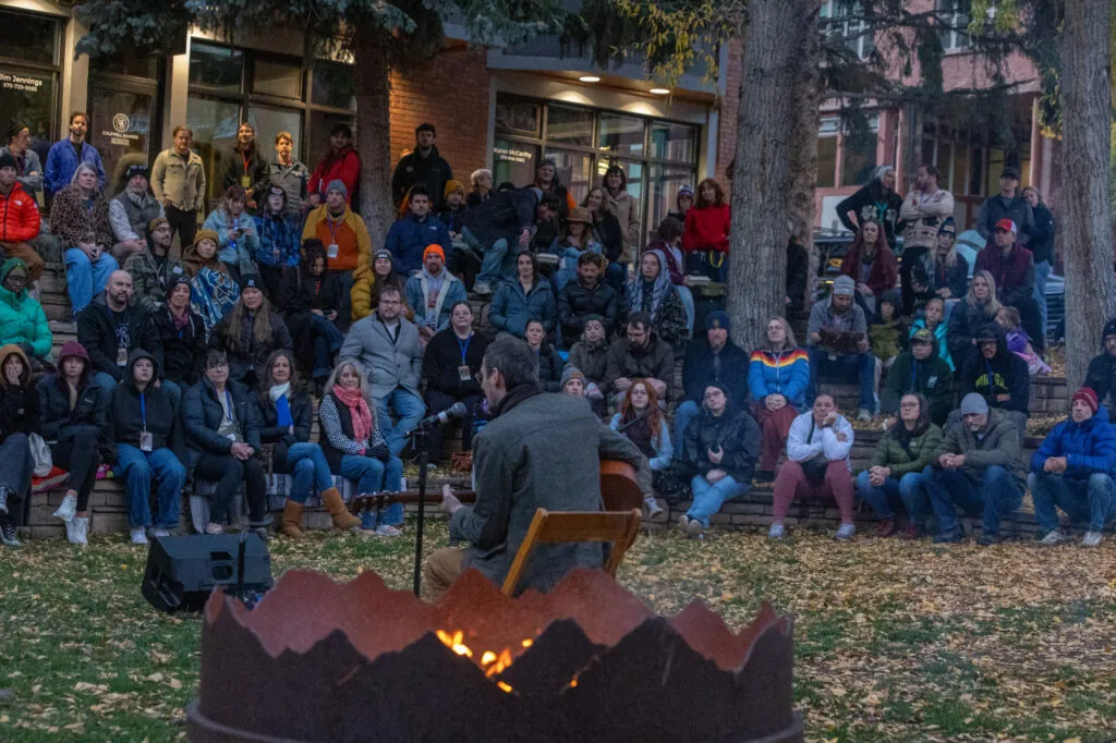 Nat Cassidy plays the guitar, back to the camera, with a crowd in the background and a bonfire in the foreground.